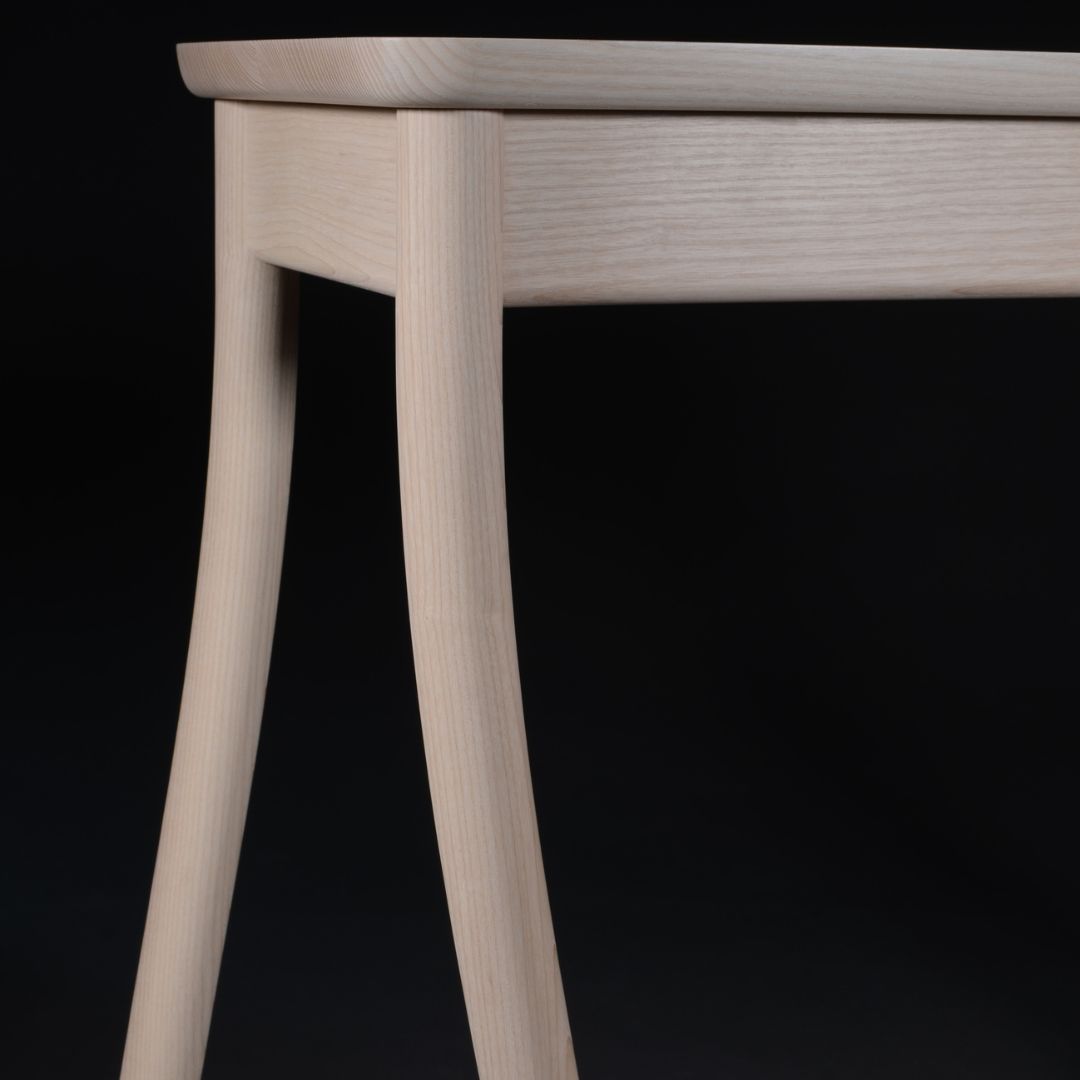 walking-table-square-2 Close-up of a light wooden stool leg and skirt against a black background, showing natural wood grain and curved legs.