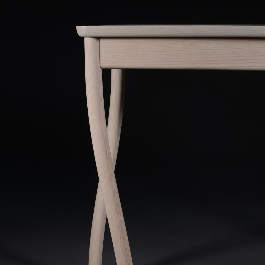 Light wooden table with crossed, sculptural legs against a black backdrop.